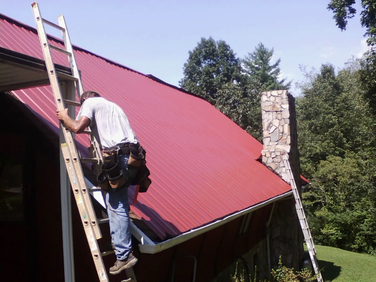 Roofer on a ladder beside a red metal roof and stone chimney — Soffit & Fascia Repair by Roofing Brothers in Killingworth
